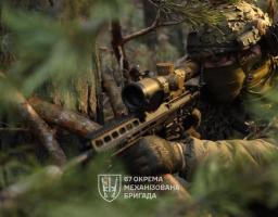 Український військовий 67-ї окремої механізованої бригади у повному бойовому спорядженні та камуфляжі прицілюється з гвинтівки з масивним оптичним прицілом, знаходячись у густих лісових хащах.
