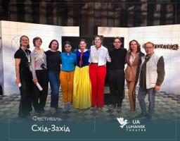 На фоні сценічних декорацій із білої плитки стоїть група з дев'яти людей, серед яких виділяється жінка в костюмі Білосніжки, а внизу розміщено написи «Фестиваль Схід-Захід» та емблему Луганського театру