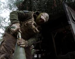 Український військовий у тактичному спорядженні та балаклаві зосереджено готує артилерійський снаряд до пострілу на фоні замаскованої бойової машини в лісистій місцевості