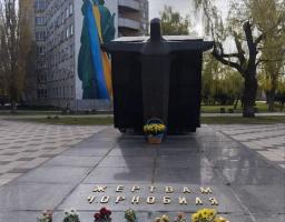 На передньому плані фотографії розташовано гранітний пам'ятник «Жертвам Чорнобиля» із покладеними біля підніжжя квітами, а на задньому плані височіє багатоповерхівка з великим муралом, де Статуя Свободи одягнена у синьо-жовтий прапор та вінок із соняшників.