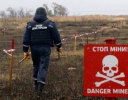 На відкритому полі спиною до глядача йде сапер у темно-синьому захисному спорядженні з написом «САПЕР ДСНС УКРАЇНИ» на спині, оглядаючи розмічену кілками та стрічками територію, а на передньому плані праворуч встановлено червону попереджувальну табличку з білим черепом і написами «СТОП МІНИ!» та «DANGER MINES!»