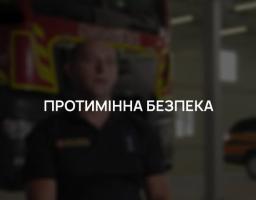 На затемненому фоні, де розмито видно чоловіка у формі рятувальника на фоні пожежної машини, по центру розміщено чіткий білий напис великими літерами «ПРОТИМІННА БЕЗПЕКА»