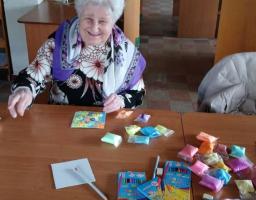 Літня жінка з білим волоссям і доброзичливою усмішкою, одягнена в чорну кофту з квітковим принтом і білу хустку з великими квітами та фіолетовим краєм, сидить за дерев'яним столом у приміщенні бібліотеки, де перед нею розкладено різноколірні пакетики з пластиліном або піском, олівці та невелика готова картинка з квітами