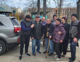 На асфальтованому майданчику біля сірого позашляховика стоїть група з восьми людей різного віку в темному осінньому одязі, які посміхаються та спілкуються між собою, а на задньому плані видно голі весняні дерева, дитячий майданчик та пошкоджену багатоповерхову будівлю.