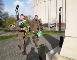 На тлі великої світло-сірої циліндричної будівлі діорами та вуличної експозиції з військовою технікою, двоє чоловіків у тактичному одязі оливкового кольору зосереджено йдуть брукованою доріжкою до білого меморіалу, тримаючи в руках великі букети жовтих і блакитних квітів, перев’язані стрічками в національних кольорах