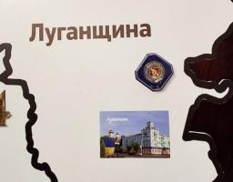 На світлій стіні розміщено частину експозиції з темним контуром карти області, де надруковано велике коричневе слово «Луганщина», а всередині закріплено синю шестикутну відзнаку з гербом та невелику горизонтальну фотографію з архітектурним пейзажем Луганська, що зображує світлу багатоповерхову будівлю зі шпилем та золоті куполи храму під ясним небом