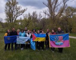 На груповій світлині, зробленій на тлі весняного лісу, зображено близько п'ятнадцяти людей, які позують на зеленій галявині, тримаючи перед собою державний прапор України та кілька прапорів територіальних громад чи організацій, зокрема великий малиново-синій прапор із гербом міста Лиман праворуч