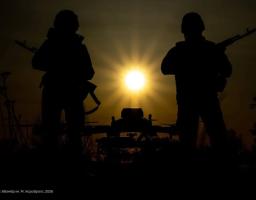 На цій драматичній світлині зображено два виразні чорні силуети українських захисників зі зброєю, які стоять по обидва боки від яскравого сонця, що сяє прямо над контурами бойового дрона, на фоні вечірнього неба з маркуванням 86-ї окремої механізованої бригади та гербом України