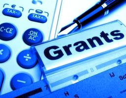 На знімку у синіх тонах зображено композицію з офісного приладдя, де на передньому плані розташована прозора папка з великим написом «Grants» (Гранти), поруч із якою лежать калькулятор із круглими кнопками та класична перова ручка.