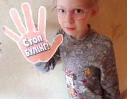 Маленька світловолоса дівчинка в рожевих окулярах і сірій сукні з Міккі Маусом стоїть біля світлої стіни, тримаючи в руці паперовий знак у формі долоні з написом «Стоп БУЛІНГ!», що символізує протест проти цькування.