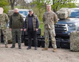 На фоні сірого пікапа та весняних дерев четверо чоловіків — двоє у військовому камуфляжі та двоє у тактичному цивільному одязі — стоять на ґрунтовій дорозі перед численними рядами чорних захисних кейсів і великих камуфляжних рюкзаків, при цьому обличчя крайнього зліва військовослужбовця приховано світлою плямою