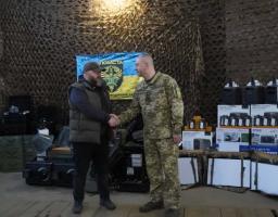 На фоні маскувальної сітки та прапора підрозділу «Помста» цивільний чоловік у жилеті та кепці тисне руку військовому в українському піксельному камуфляжі, стоячи серед численних коробок із портативними зарядними станціями Bluetti, акумуляторами та іншим технічним обладнанням.