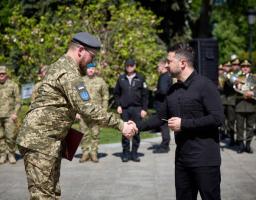 Президент України Володимир Зеленський тисне руку військовослужбовцю та вручає йому нагороду під час урочистої церемонії просто неба на фоні інших військових та дерев