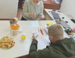 У світлому кабінеті за білим столом, заставленим ноутбуками, художнім приладдям та тарілкою з рогаликами, жінка в окулярах і світлому светрі натхненно демонструє чоловікові в оливковій куртці процес розпису білого яйця пензлем