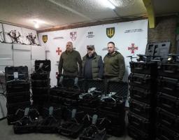 У приміщенні складу на фоні білого банера з офіційною символікою українських військових підрозділів троє чоловіків у тактичному одязі стоять в оточенні великої партії допомоги, що складається з численних квадрокоптерів у відкритих чорних кейсах та високих стопок білих коробок із технікою