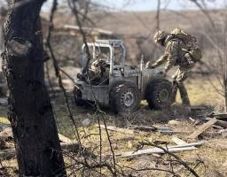 На сонячній відкритій місцевості серед сухої трави та залишків деревини солдат у камуфляжі та повному тактичному спорядженні нахиляється над невеликою чотириколісною роботизованою платформою, завантаженою речами, в той час як на лівому передньому плані височіє темний стовбур дерева