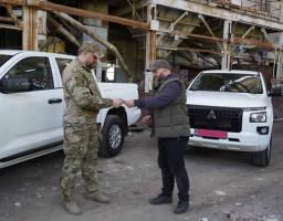 У промисловій зоні на фоні металевих конструкцій чоловік у темному жилеті та кепці передає ключі від нового білого пікапа Mitsubishi L200 військовослужбовцю у камуфляжі, а поруч стоїть ще один аналогічний автомобіль, що підкреслює момент передачі техніки для потреб оборони