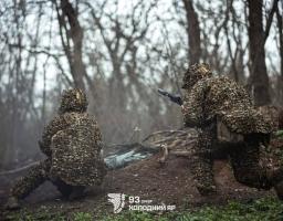 Двоє бійців 93-ї бригади «Холодний Яр» у густому маскувальному одязі типу «гіллі», що повністю приховує силуети, тримають зброю напоготові серед тонких стовбурів дерев у лісі