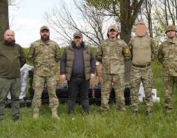 Шестеро чоловіків, четверо з яких одягнені в український камуфляж, а двоє — у тактичний цивільний одяг, стоять у ряд на зеленій галявині перед стопками чорних захисних кейсів і коробок із обладнанням на фоні високих дерев, причому обличчя одного з військовослужбовців розмите