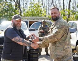 На сонячній галявині на фоні білих пікапів бородатий чоловік у цивільному жилеті та кепці з татуйованими руками передає ключі та тисне руку військовослужбовцю у камуфляжній формі, у той час як на задньому плані стоїть солдат зі зброєю, а поруч на землі складено чорні кейси з обладнанням