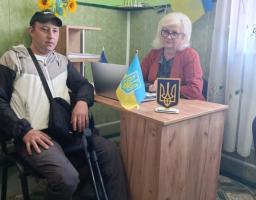 У кабінеті з зеленими стінами, декорованому державними символами України, за робочим столом із ноутбуком сидить жінка в червоному піджаку, а поруч на стільці — чоловік у спортивній кепці, який тримає металеву милицю.