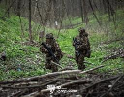 На фото двоє українських військових у маскувальних костюмах «гіллі», що імітують сухе листя та гілки, обережно просуваються лісистою місцевістю з гвинтівками в руках, а внизу зображення розміщено емблему та напис «93 ОМБр ХОЛОДНИЙ ЯР».