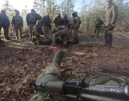 На знімку, зробленому від першої особи з позиції лежачи, видно руки військового з автоматом на фоні групи бійців у камуфляжі, які зібралися в осінньому лісі навколо пораненого побратима для надання допомоги