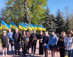 Близько одинадцяти людей різного віку стоять у шеренгу на брукованій доріжці. Майже у кожного на грудях прикріплена червона квітка маку — символ пам'яті. У руках деякі учасники тримають квіти (жовті та червоні гвоздики, синьо-жовті букети)