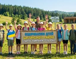 Група радісних дітей та дорослий чоловік махають руками на фоні мальовничого гірського пейзажу, тримаючи перед собою великий банер про старт кампанії з оздоровлення дітей Щастинського району 2026 року