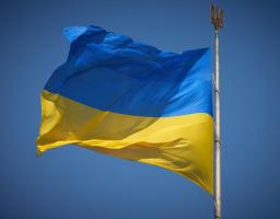 Державний прапор України з двох рівновеликих горизонтальних смуг синього і жовтого кольорів майорить на вітрі на високому металевому флагштоку, верхівку якого прикрашає золотистий тризуб на тлі чистого безхмарного неба