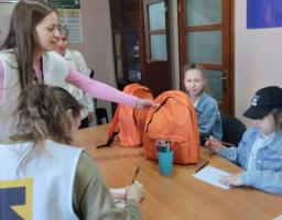 Зліва стоять двоє дівчат-волонтерок. Одна з них, з довгим русявим волоссям, одягнена у світлий жилет поверх рожевої кофти. Вона простягає руку до яскраво-помаранчевого рюкзака, що стоїть на столі. Інша волонтерка, яку видно зі спини, також у білому жилеті з жовто-чорним логотипом, щось занотовує на папері
