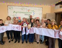 Люди стоять у два ряди. Перед собою вони тримають довгі білі рушники з червоно-чорною вишивкою, що тягнуться вздовж усієї групи. Деякі учасники тримають у руках невеликі книжки або брошури