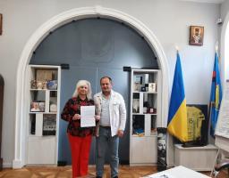 Жінка у яскравих червоних штанах і квітчастій блузі та чоловік у світлому піджаку стоять у кабінеті на фоні великої декоративної арки, полиць із пам'ятними речами та державного прапора України, разом тримаючи перед собою офіційний документ.