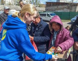 Молода жінка-волонтерка зі світлим волоссям, зібраним у хвіст, стоїть спиною до глядача ліворуч. Вона одягнена у яскраво-синю толстовку (худі) з жовто-зеленим логотипом на спині. Вона з посмішкою простягає відкриту картонну коробку з різнокольоровими дрібними предметами (схоже на цукерки в обгортках) людям перед нею