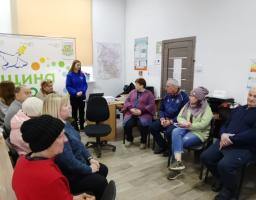 У світлому кабінеті група людей старшого віку сидить півколом і уважно слухає дівчину в синій кофті, яка стоїть біля ноутбука та проєктора на фоні інформаційного стенда з логотипом у вигляді голуба