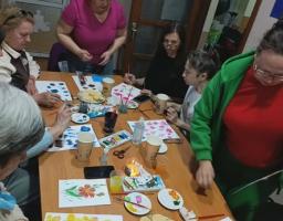 Група жінок різного віку зосереджено займається малюванням за довгим дерев'яним столом, на якому серед розкладених фарб, пензлів та склянок лежать готові малюнки з яскравими квітами, чисті аркуші та тарілки з частуванням