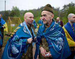 На передньому плані світлини під відкритим похмурим небом зображено двох усміхнених чоловіків у військовій формі, загорнутих у сині брендовані прапори з українською символікою та написами, причому ліворуч стоїть лисий військовий середнього віку, а праворуч — старший чоловік у зеленій пілотці з пачкою сигарет у руці, тоді як на задньому плані видно інших побратимів із синьо-жовтими прапорами на тлі зеленого лісу.