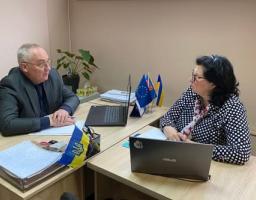 У робочому кабінеті за двома зсунутими дерев'яними столами, де стоять два відкриті ноутбуки, стоси документів та маленькі прапори України і Євросоюзу, сидить літній сивіючий чоловік у сірому піджаку та окулярах, який дивиться і розмовляє з чорнявою жінкою в окулярах та чорно-білій блузці в горошок, що сидить навпроти нього