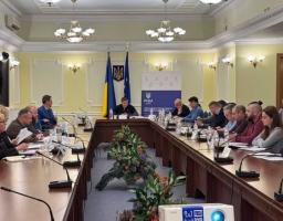 У світлій офіційній залі за великим П-подібним дерев'яним столом з настільними мікрофонами, документами та квітковими композиціями в центрі проходять збори або засідання комітету, де понад двадцять учасників у цивільному одязі сидять уздовж столів, а в торці зали під прапорами України та Європейського Союзу і гербом на стіні головує чоловік у темному одязі.