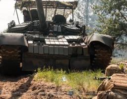 На лісовій ґрунтовій дорозі великим планом спереду зображено український танк, оснащений динамічним захистом та антидроновою сіткою-навісом, з люків якого виглядають члени екіпажу, тоді як на передньому плані праворуч спиною до глядача в окопі лежить військовий у касці та бронежилеті зі зброєю, а ліворуч удалині стоять ще двоє бійців.