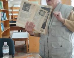 У приміщенні бібліотеки з дерев'яними книжковими стелажами та підвісною стелею сивочола жінка у світло-сірому жилеті з накладними кишенями стоїть біля столу і демонструє розгорнуту книгу з ілюстраціями круглих візерунків та текстом, у той час як на передньому плані на дерев'яній стільниці розкладено книги, прозору пластикову пляшку з водою, окуляри та мотки ниток і шнурів для рукоділля
