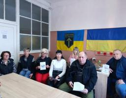 На світлині зображено групу з семи людей, які сидять за столом у кабінеті на тлі стіни з двома українськими прапорами (один із яких містить емблему з написом «SEMPER PRIMUM» та стрілкою вгору), при цьому дехто з присутніх тримає в руках інформаційні буклети та папери