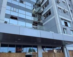 На світлині під кутом знизу вгору зображено багатоповерхову сучасну будівлю сірого кольору з великим скляним фасадом, на якому видно численні вибиті вікна та пошкодження, при цьому весь перший поверх та вхідна зона закриті захисними дерев'яними ОСБ-плитами, а на вищих поверхах помітні зовнішні блоки кондиціонерів