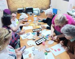 На світлині згори зафіксовано творче заняття, під час якого семеро жінок різного віку сидять за довгим дерев'яним столом у кабінеті на тлі великої старовинної карти Європи і розписують фарбами камінці та папір, використовуючи художні пензлі, пластикові палітри, стаканчики з водою та олівці.