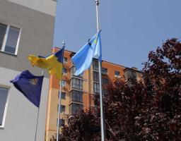 На світлині знизу вгору на тлі чистого синього неба зображено три прапори на високих металевих флагштоках — темно-синій прапор із круглою емблемою ліворуч, державний синьо-жовтий прапор України по центру та блакитний прапор із золотою кримськотатарською тамгою праворуч, при цьому на знак жалоби до верхньої частини кримськотатарського стяга прикріплено чорну стрічку, а на задньому плані видно частину сучасної житлової будівлі та густу крону дерева з темно-бордовим листям.