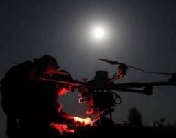 На нічній світлині зображено темні силуети двох військових у шоломах, які при світлі червоного тактичного ліхтаря налаштовують великий багатороторний безпілотник (гексакоптер чи октокоптер) на тлі повного місяця, що яскраво світить у нічному небі