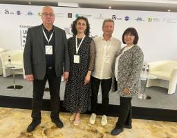 Чотири учасники конференції — двоє чоловіків та дві жінки з бейджами на шиї — стоять у ряд на сцені офіційного заходу на тлі великого білого банера з логотипами та білих крісел.
