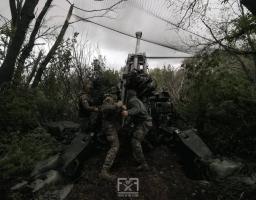Військові в камуфляжі обслуговують велику артилерійську установку, що замаскована сіткою серед густих дерев та кущів під похмурим небом
