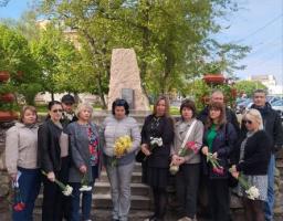 На світлині зображено групу з одинадцяти людей, які стоять півколом на вулиці біля кам'яного пам'ятного знака в оточенні зелених дерев, при цьому майже всі присутні тримають у руках весняні квіти, такі як нарциси, тюльпани та гвоздики