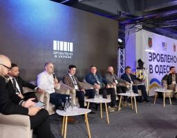 Група чоловіків у костюмах сидить у кріслах на сцені під час офіційного заходу на тлі великих екранів із написами «Зроблено в Україні» та «Зроблено в Одесі»